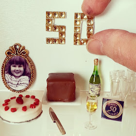 Modern dolls' house miniature 50th birthday scene with cake, champagne and a gold diamante '50' on the wall.