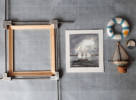Flat lay of a 1/12 scale miniature frame in a jig, next to a grey watercolour of a boat, on a piece of card, next to a 1/12 scale model yacht, round picture of a yacht and a blue and white lifebuoy.