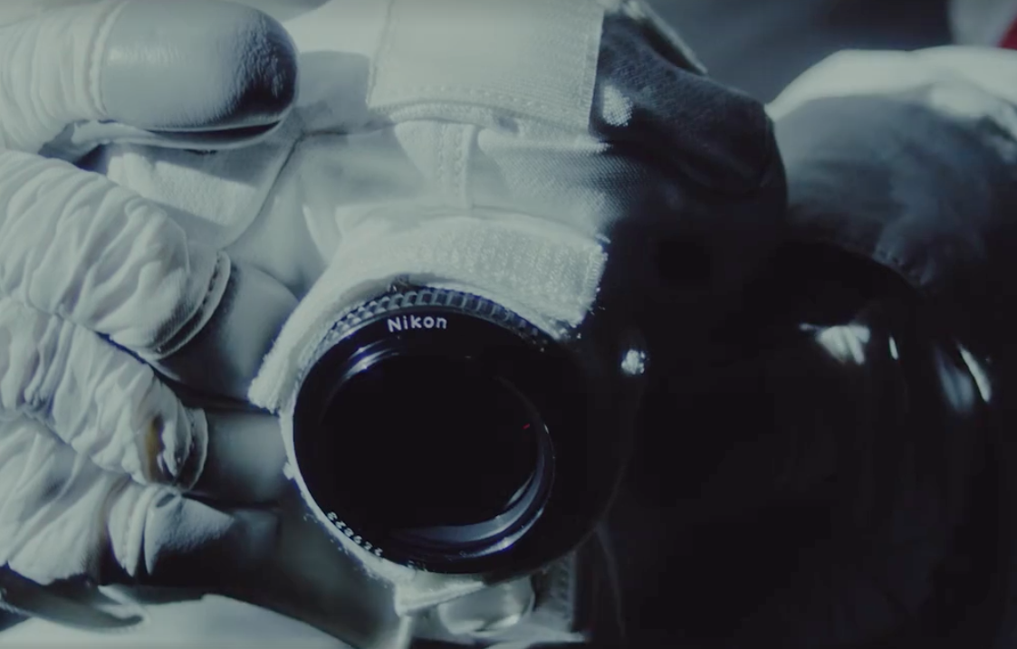 Nikon Kamera und Objektiv in den Händen eines Astronauten - Man sieht die Handschuhe und die eingepackte Kamera in weißem Thermostoff auf dem Objektiv steht Nikon