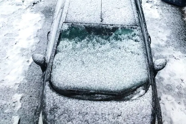 A car in a drive way covered in a light layer of snow which needs removing for driving safely