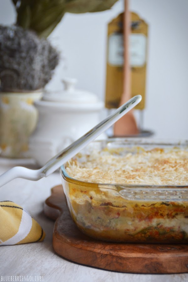 Lasaña de berenjenas en fuente de cristal sobre tabla de madera, con ingredientes de fondo, hierbas aromáticas, laurel seco, 