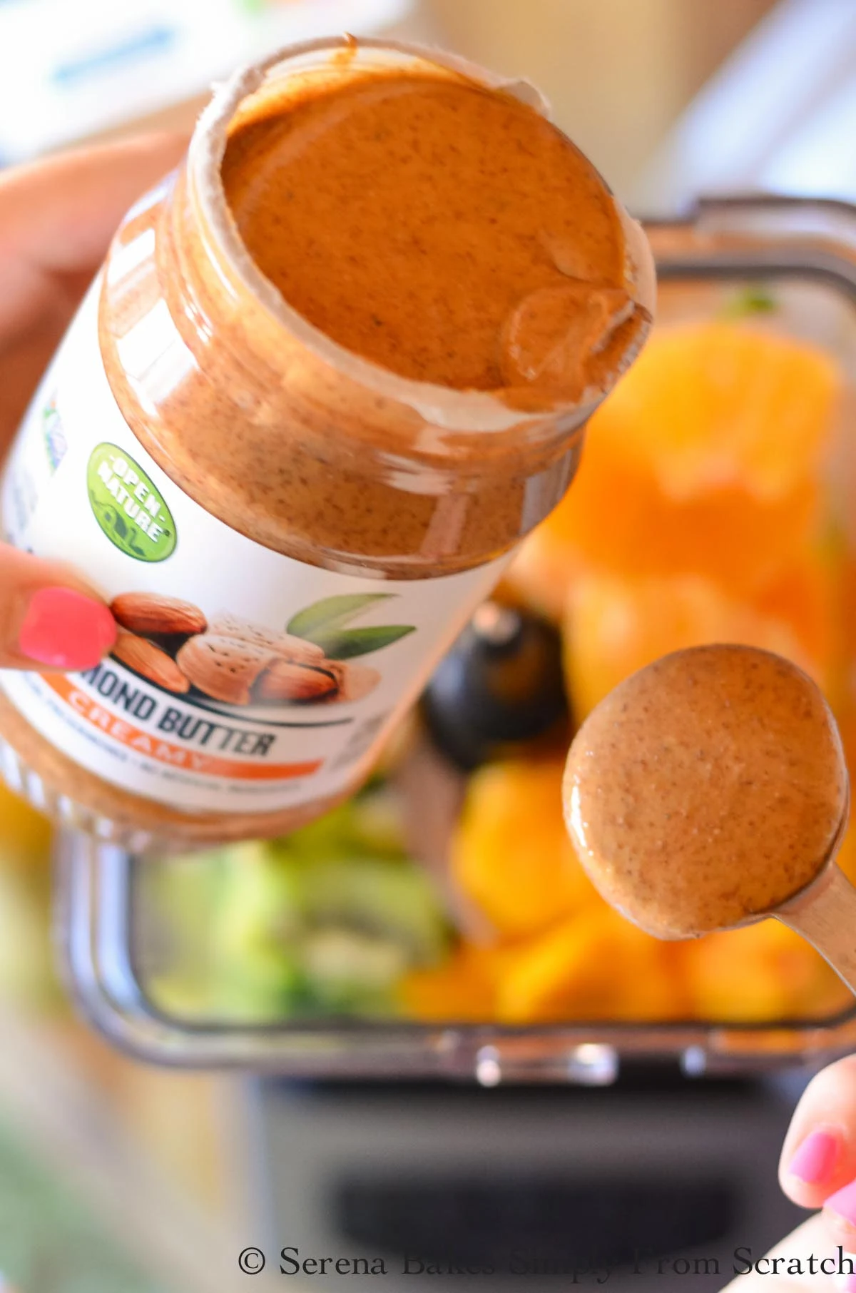 A container of almond butter and a tablespoon filled with almond butter over blender.