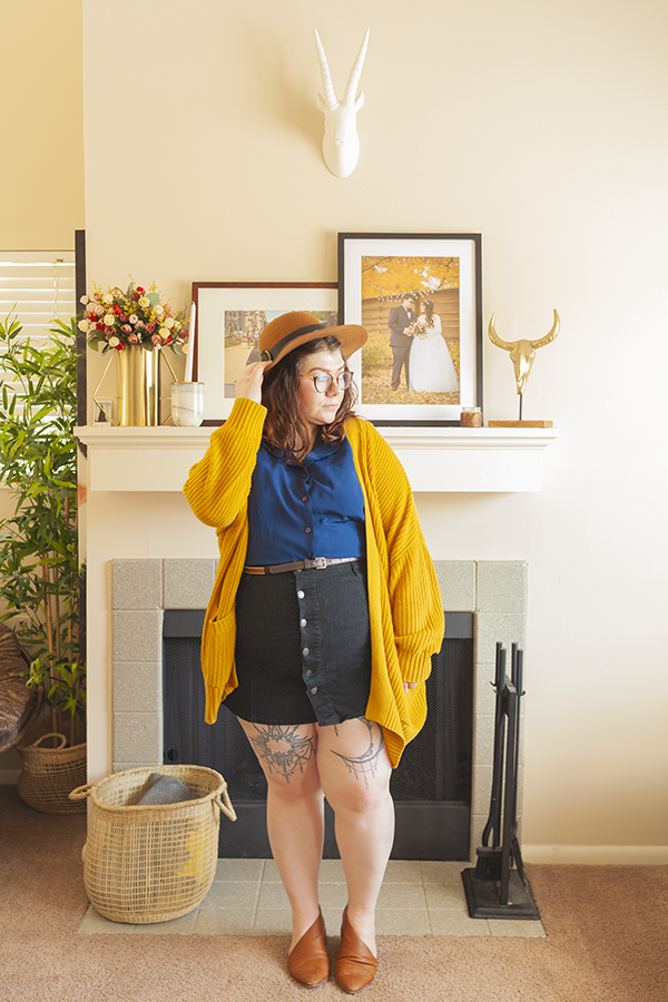 An outfit consisting of a brown panama hat, mustard yellow chunky cardigan, navy blue blouse tucked into a black button down skirt and brown d'orsay flats.