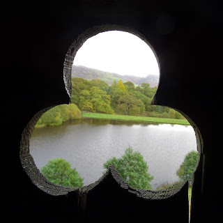 A photo showing a view through a hole in the shape of a spade in a wooden door in the eastern tower of Morton Castle.  It shows some trees and a body of water.  Photograph by Kevin Nosferatu for the Skulferatu Project.