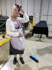 A woman dressed as char lady poses with her broom in front of an empty table.