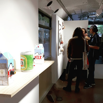 Shelf in a gallery holding a collection of milk cartons and beer cans made out of cardboard. Behind it a group of people look at some assemblage pieces on the wall.