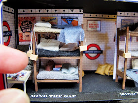 Miniature scene of a three-tier bunk in an underground shelter set up in a 1940s tube station. with fingers for scale.