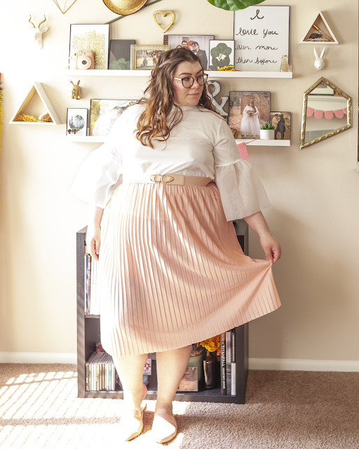 An outfit consisting of a high neck white blouse with sheer trumpet sleeves at the elbow tucked into a pleated pastel pink midi skirt and muted pink pointed toe mules.