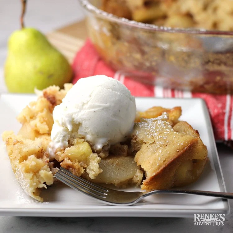 Warm Pear Cobbler by Renee's Kitchen Adventures on a white plate with a scoop of vanilla ice cream and a fork with a fresh pear in the background on red and white linens