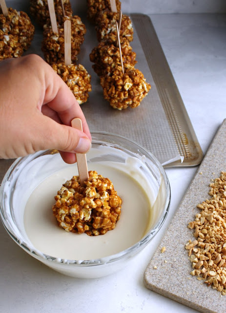 dipping caramel apple popcorn balls in white chocolate with tray of chopped peanuts nearby