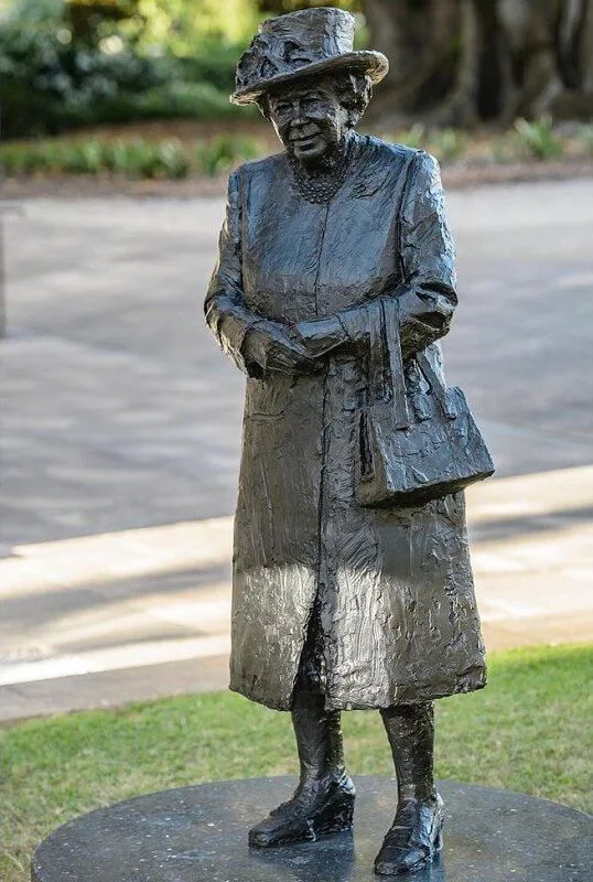 The sculpture depicts Queen Elizabeth in a coat and hat carrying her trademark Launer handbag. diamond silver brooch and pink dress