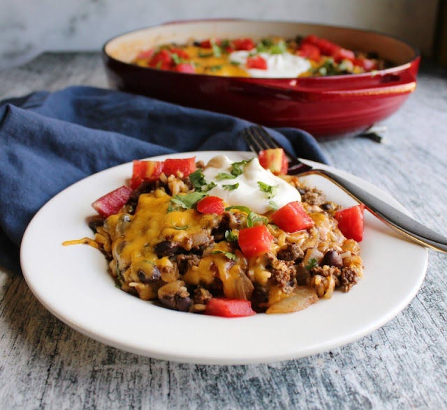One pan taco rice skillet is a quick and easy way to get dinner on the table. It has maximum flavor with minimal dirty dishes! close up of a plate loaded with one pan taco rice skillet meal topped with melted cheese, tomatoes, sour cream and cilantro with red cast iron pan in background