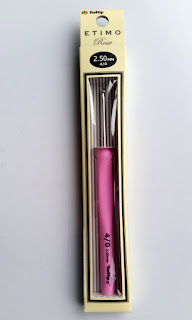 A pink handled, silver aluminium crochet hook is displayed in its packaging - a cardboard sleeve or narrow box with a transparent cellophane window. The hook has its size printed in black lettering on its handle and also printed on the box.
