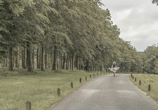 girl in white dress walks along road bordered by tall green trees