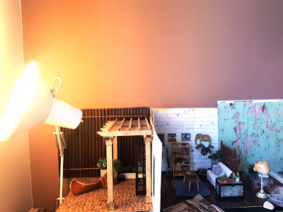 One twelfth scale modern miniature scene of a courtyard and a lounge with french doors leading to it. To the left of the photo is an angelpoise lamp, trained at the wall next to the courtyard to provide some false sunlight.