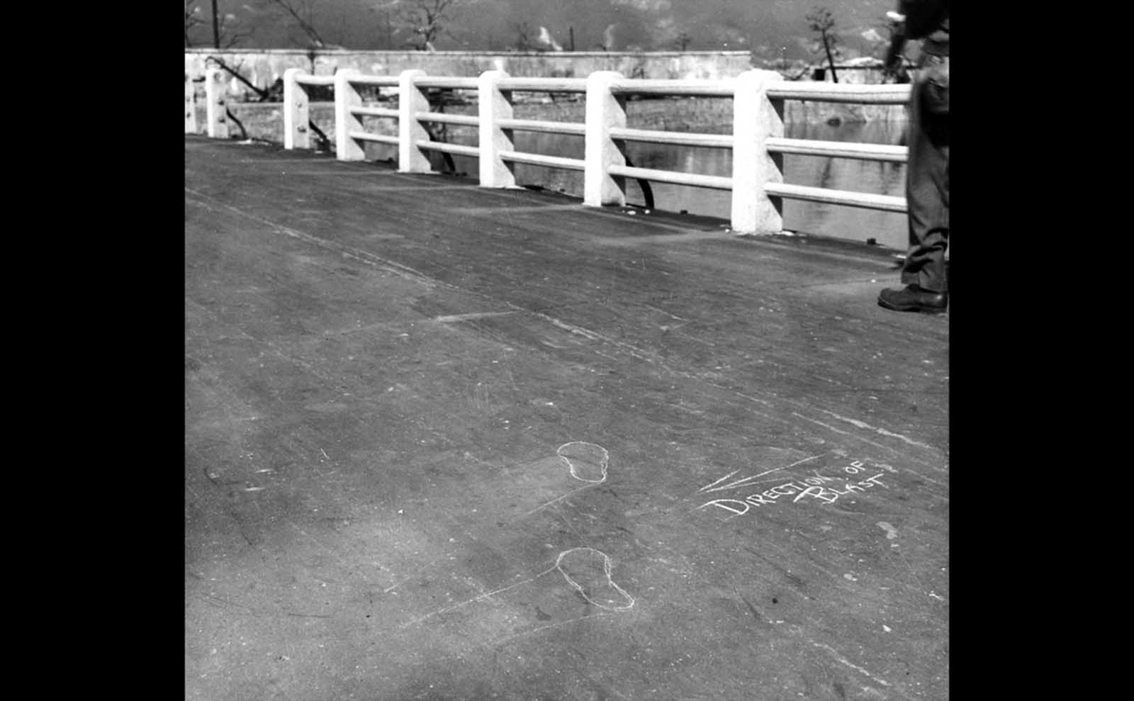 “Direction of blast” chalk marks and outlines of the feet of a victim caught in the explosion. The intense heat of the initial flash of the detonation seared every nearby surface, leaving inverted “shadows,” like those seen on this bridge left by the railings and by a person who had been standing in this place.