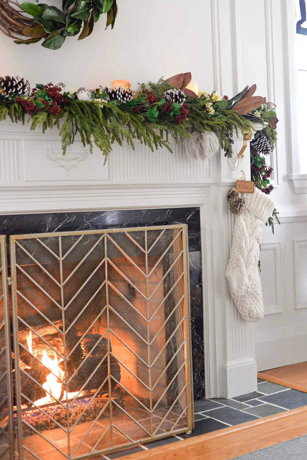 a faux christmas fireplace mantel garland with red seeded eucalyptus, a cedar garland and magnolia leaves. A vine and magnolia asymmetrical wreath with satin ribbon and metal bells. A brass chevron fireplace screen.