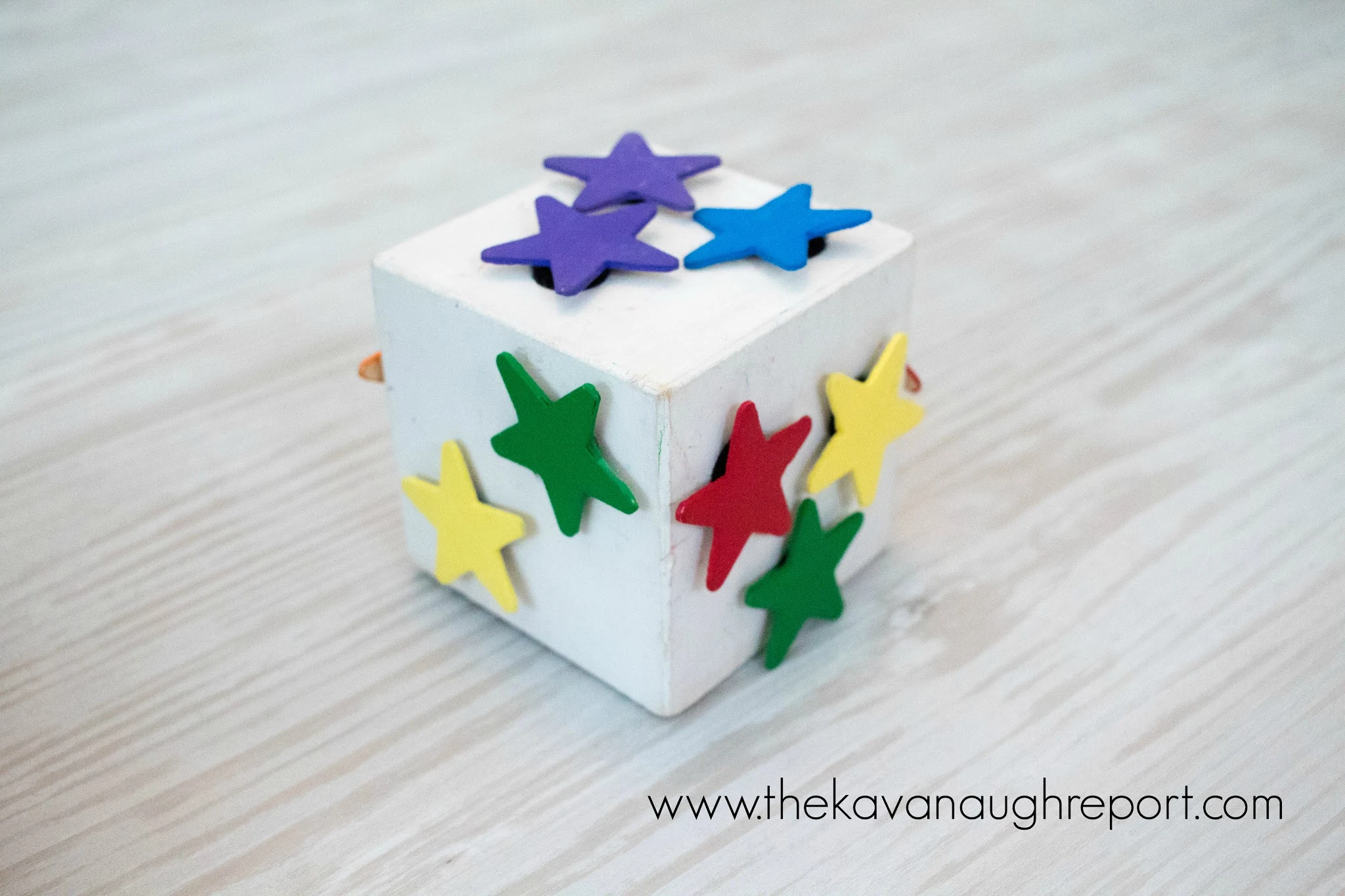 A DIY wooden block toy sits on the floor. It is painted white and covered with colorful wooden stars waiting for a toddler to play with it.