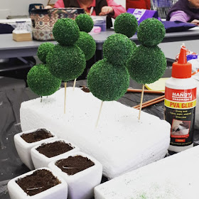 Six one-twelfth scale topiaries skewred to a piece of styrofoam next to four planters filled with miniature soil.