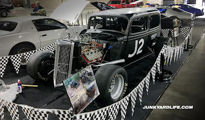 Black 1934 Ford race car restored after rescue from woods is displayed at World of Wheels car show behind checkered flag that line the area around car.