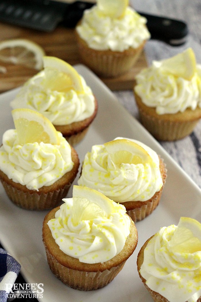 Easy Lemon Cupcakes by Renee's Kitchen Adventures on a white platter garnished with lemon wedges and yellow sanding sugar with a few cupcakes off the platter to the side and a knife and cutting board with lemons in the back