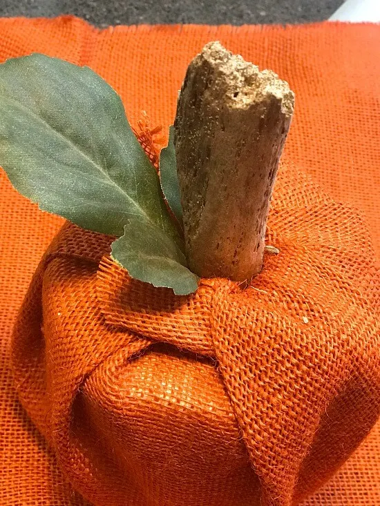 Stick and leaf sticking out of top of orange burlap toilet paper cover.