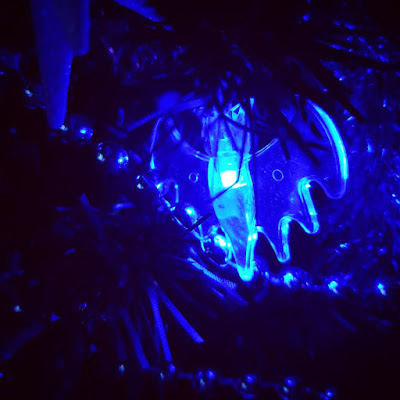 The black Christmas tree is illuminated by a light that appears blue; in the centre is a lit bat LED light, it is brightest in the middle, so bright it is almost white in the photograph. The rest of the tree is very dark, much is black. In real life, the bat glows a more purple-y colour.