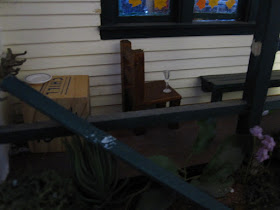 Early-morning shot of a one-twelfth scale model school veranda, with a broken railing, a table with an empty plate, and a school chair with an empty wine glass on its seat.