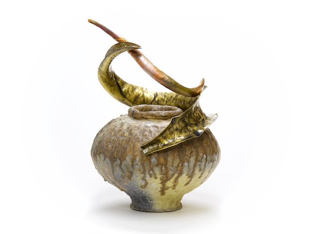 Rounded wood-fired porcelain jar with multi-coloured wood ash-glaze deposits, rests on a small foot-ring, with curved sculptural brass and bronze forms wrapped around the shoulders and top opening, front view.