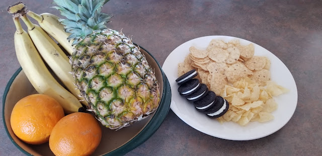 Bananas, oranges, a pineapple in a bowl next to a plate with chips and cookies.