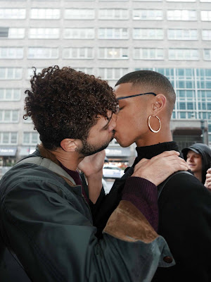 ID: two people, one on the left with dark skin, dark curly hair, and a beard and one on the right with dark skin, close-cropped black hair, glasses, and a large hoop earring, share a kiss.