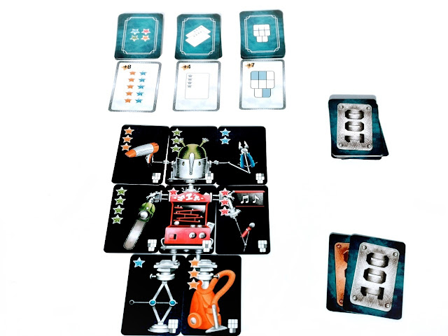 gra karciana Robot X, na zdjęciu rozłożone karty jednego z graczy, na środku ukończony robot, obok leża karty pieniężne, nad robotem widać stosy kart bonusowych do dobierania, 