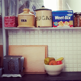 1/12 scale modern miniature scene of a kitchen bench with a fold-down toaster, chopping board and bowl of fruit on it. On the shelf above are two containers of tea, a cannister of sugar, a packet of Weet-Bix and a half-used jar of jam.