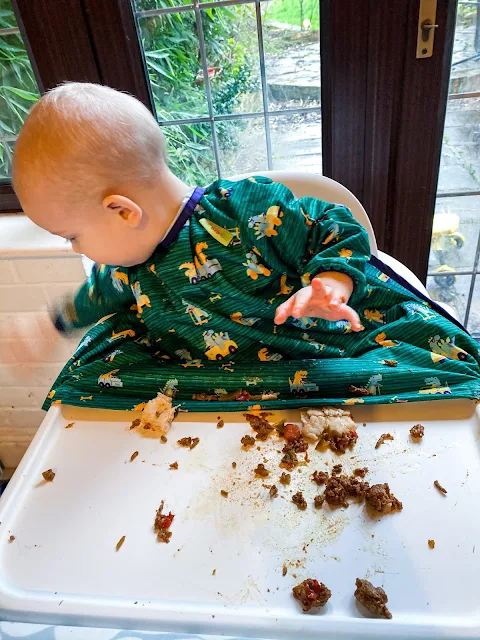 A photo of a baby in a highchair with food all over the tray. He is looking at the floor on the side of the highchair and waving his hand fast (it is blurred)
