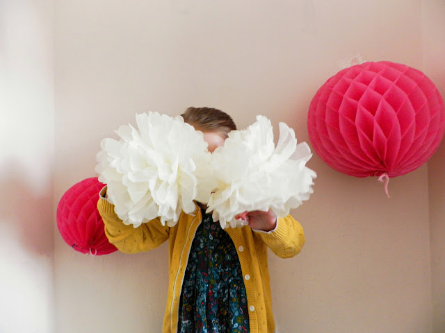 A blonde haired girl wearing a green floral patterned dress and yellow cardigan is holding two white fluffy paper balls in front of her face with two large pink paper ball decorations on a cream wall behind her. She is stood under a pink banner saying Happy Birthday.