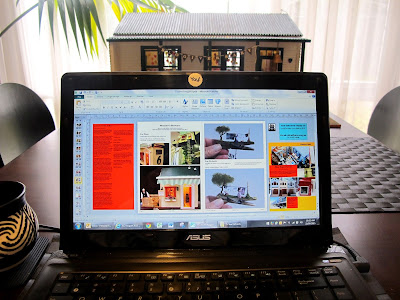 Laptop computer showing a magazine design in progress, on a dining table. AT the other end of the table is a dolls' house miniature school building.