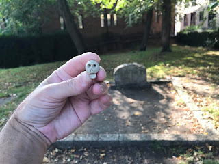 A picture showing a hand holding a small, ceramic skull with the grave of Dick Turpin in the background.  Photo by Kevin Nosferatu for the Skulferatu Project.