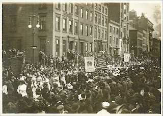 A black and white photograph of a parade moving through a crowded street.