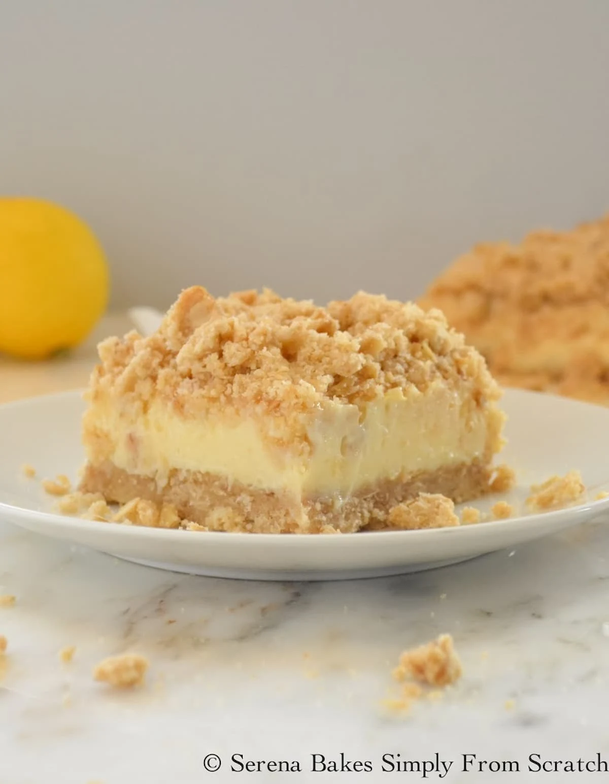 Sliced Lemon Cheesecake Crumb Bars on a white plate with a pan of Lemon Cheesecake Crumb Bars in the background.