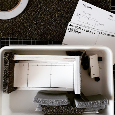 Aerial view of a plastic container holding a half-made one twelfth modern miniature sofa, with various components to finish it surrounding the container: double-sided tape, spare fabric, foam and pins for feet.