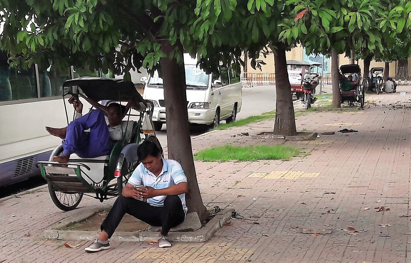 Two tuk tuk drivers resting under a tree next to their tuk tuk.