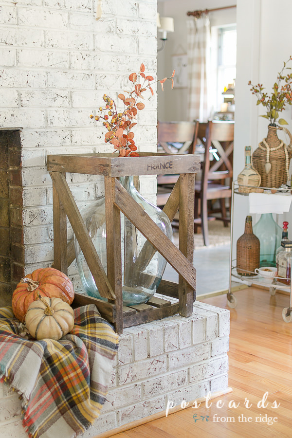 large blue glass bottle in wooden crate, plaid blanket scarf with faux pumpkins, on a painted brick hearth