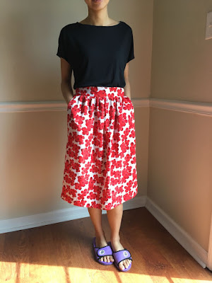 A person wearing a black top and red/white floral skirt in purple slippers.
