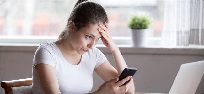 امرأة شابة منزعجة تحمل هاتفًا ذكيًا وتنظر إلى شاشة الكمبيوتر.