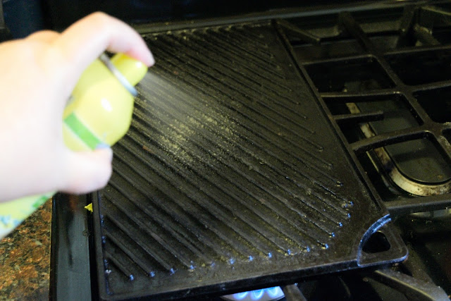A cast iron grill pan, on the stove, with cooking spray being sprayed on it.  