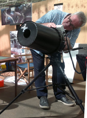 A vortex machine is poised upon a tripod.  It looks like a giant camera lens with a hole in the centre but it is created with a plastic bucket!  The operator is aiming the barrel to shoot a burst of air.