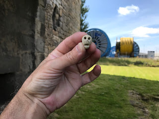 A photo of Skulferatu #42 being held up with walls of castle keep on left hand side and industrial scenery from Rosyth Dockyard behind.  Photo by Kevin Nosferatu for the Skulferatu Project