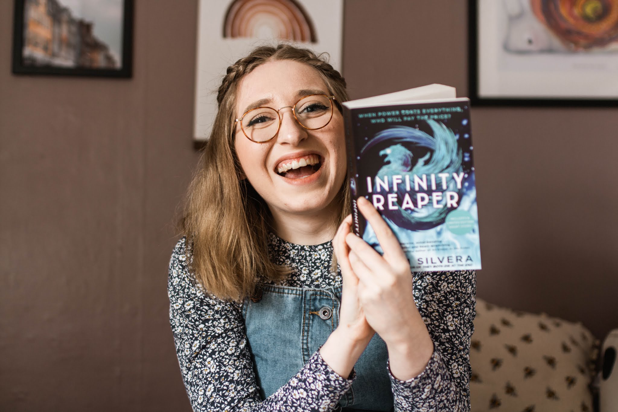 blonde girl with glasses smiling wearing blue and holding blue book infinity reaper by adam silvera