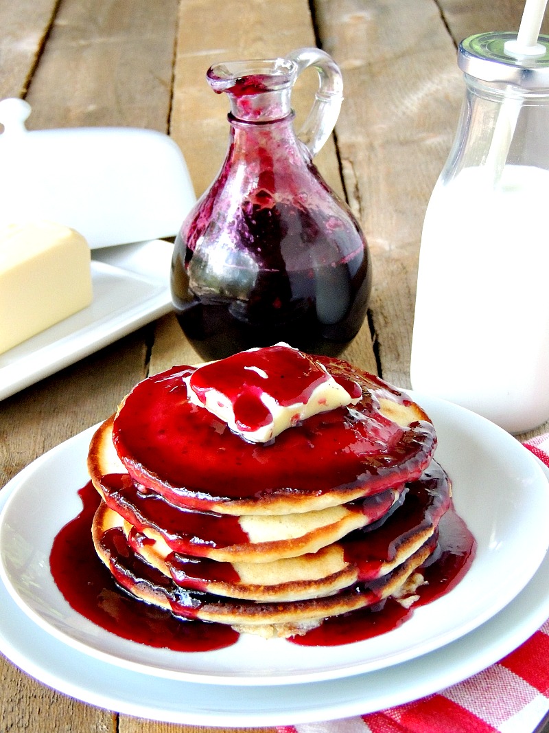Glass bottle with Homemade Blueberry Syrup in it on a wooden table. Butter dish, glass of milk, and pancakes with butter and blueberry syrup on a white plate with it.
