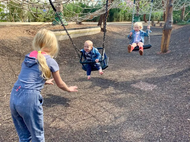 Older child pushing baby on swing with a 3 year old behind also on a swing